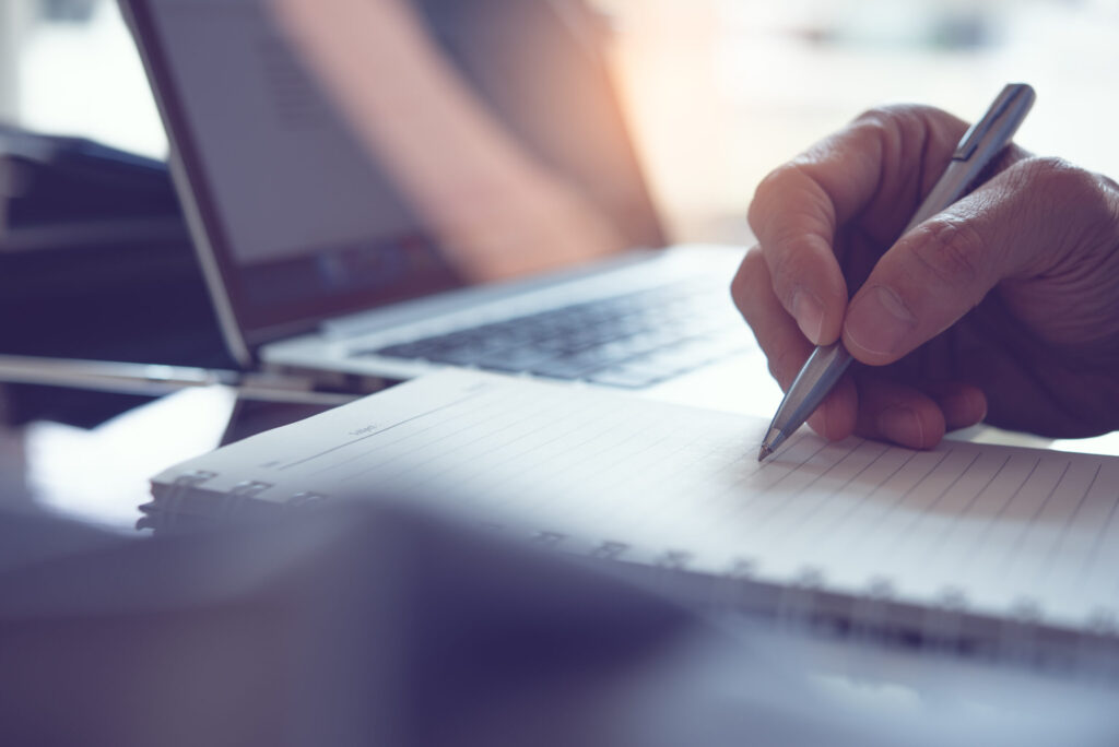 Business man hand write his work plan on paper notebook with a pen while working in modern office. Man student taking note while learning online course via laptop computer, close up, elearning concept